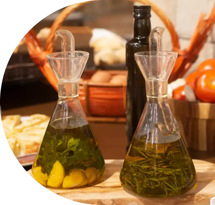 Glazen flessen met kruiden en knoflook in olie staan op een houten plank. Daarachter staat een mand met brood, een fles en een schaal met tomaten, wat een gezellig keukentafereel oplevert.